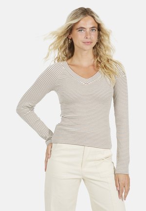 Blondynka o falowanych włosach, ubrana w beżowo-białą koszulę w paski z długim rękawem i dekoltem w serek oraz jasnobeżowe spodnie, stoi z jedną ręką na biodrze.