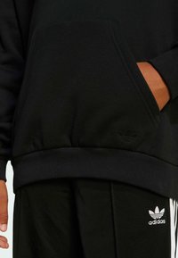 Černá mikina s klokaní kapsou, žebrovanými manžetami a lemem. Disponuje diskrétním černým logem a je kombinována s odpovídajícími černými kalhotami.