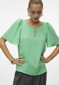 Blusa verde con maniche corte a sbuffo, scollatura a chiave e tessuto testurizzato. Abbinata a pantaloni grigi scuri.
