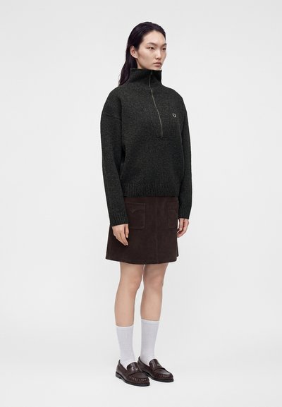 Femme portant un pull vert foncé avec fermeture éclair sur la moitié, une jupe marron foncé avec des poches avant, des chaussettes blanches et des mocassins marron, debout devant un fond uni.
