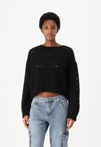 Svart strikket kort genser med et åpenarbeidet design, rund hals og lange ermer, kombinert med lyseblå cargo-jeans.