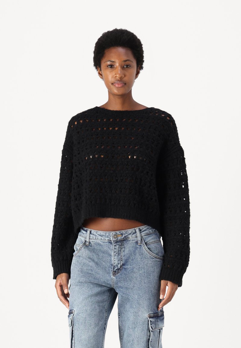 Svart strikket kort genser med et åpenarbeidet design, rund hals og lange ermer, kombinert med lyseblå cargo-jeans.