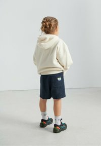 Sweat à capuche en polaire crème avec cordon de serrage à la capuche, associé à un short bleu marine effet serviette. Baskets noires et vertes, chaussettes blanches avec détails logo.