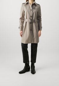 Beige trench coat with a belted waist, button front, shoulder epaulettes, and a smooth texture. Paired with black denim and ankle boots.