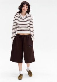 Top cropped com riscas horizontais castanhas e brancas, combinado com calções castanhos folgados e mocassins castanhos, com meias brancas até ao tornozelo visíveis.