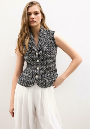 Γυναίκα που φοράει αμάνικο ασπρόμαυρο υφασμάτινο γιλέκο με κουμπιά από πέρλες και λευκό ψηλόμεσο πλισέ παντελόνι, στέκεται μπροστά σε απλό φόντο.