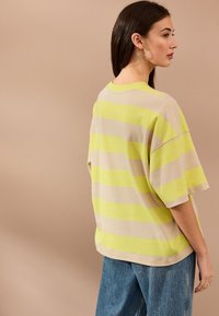 Nadrozmerné béžové tričko s krátkym rukávom so žltými a svetlosivými horizontálnymi pruhmi, v kombinácii s modrými džínsami. Hladká textúra látky.