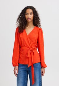 Blouse rouge avec un motif texturé, manches bouffantes et une ceinture nouée à la taille. Associée à un jean en denim bleu.