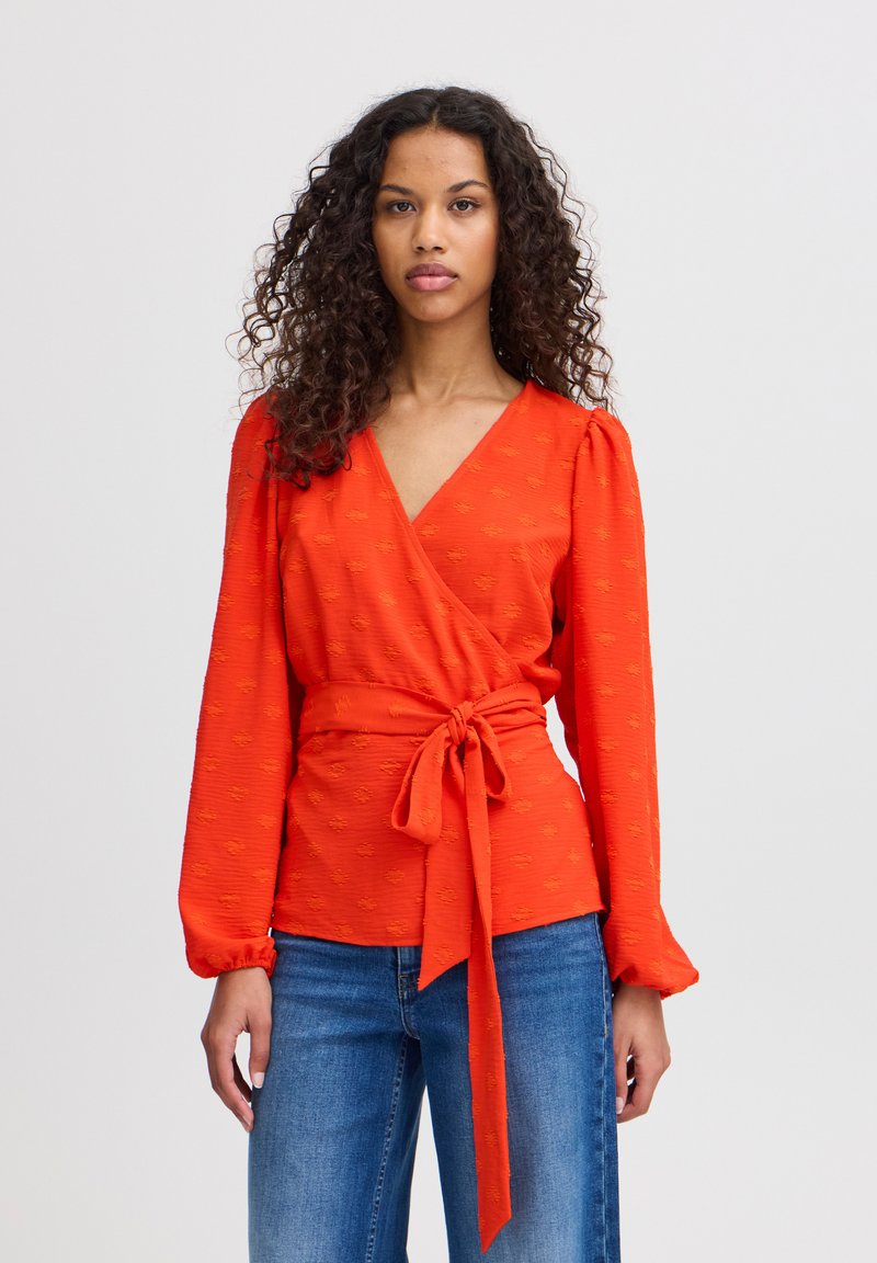 Blouse rouge avec un motif texturé, manches bouffantes et une ceinture nouée à la taille. Associée à un jean en denim bleu.