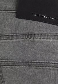 Šedá džínovina s detailem šití a koženou etiketou s vyraženým nápisem "TRUE RELIGION." Detailní záběr zaměřený na texturu a barvu.