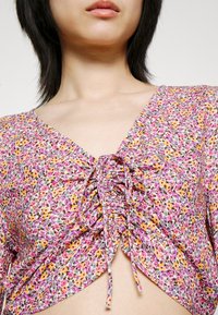 Femme portant un haut à col en V fleuri avec un lien froncé à l'avant, de petites fleurs roses, jaunes et violettes sur fond blanc.