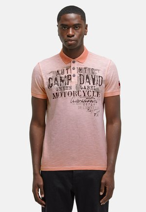 Polo à manches courtes en rose clair avec un col contrasté, arborant un imprimé graphique en lettres foncées. Fabriqué en tissu doux et texturé.