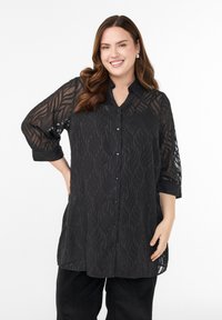 Usmievavá žena s vlnitými vlasmi, ktorá má na sebe polopriesvitnú čiernu vzorovanú blúzku na gombíky a čierne nohavice, pózujúca s jednou rukou na bokoch.