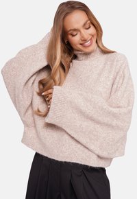 Uśmiechnięta kobieta z długimi falującymi włosami nosi oversize'owy beżowy sweter z golfem i czarne plisowane spodnie, pozując z jedną ręką na głowie.