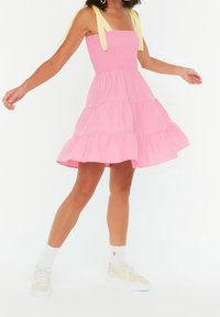 Rosa, gestuftes Sommerkleid mit gerafftem Oberteil und gelben Trägern. Kombiniert mit weißen Socken und hellen Sneakern. Glatte Textur, lässiges Design.