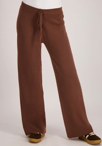 Bruine wijde broek met een trekkoord in de taille, gemaakt van zachte stof. Ze hebben een gladde textuur en een vloeiende silhouet, gecombineerd met donkere schoenen.