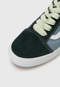 Baskets avec une tige en daim vert foncé, des accents textiles bleu clair, des coutures blanches et des lacets vert pâle. Semelle en caoutchouc blanche visible en dessous.