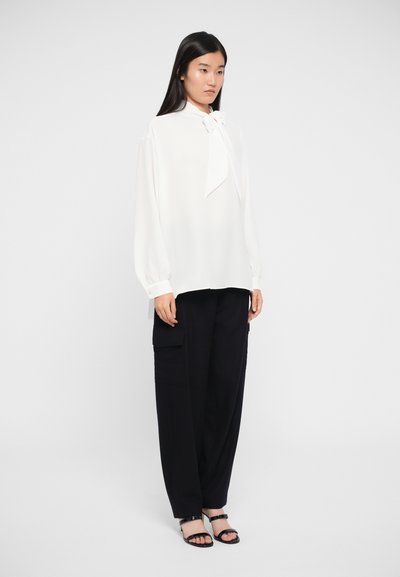 White blouse with a bow tie detail at the collar, long sleeves, paired with black cargo pants and black strappy sandals.
