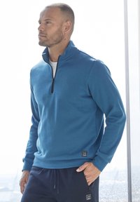 Blue zip-neck pullover made of soft fabric with a relaxed fit, featuring a black logo patch at the hem and a smooth texture.