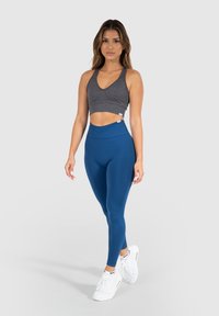 Grau geschnittenes Sport-BH mit tiefem V-Ausschnitt, kombiniert mit hochtaillierten blauen Leggings. Aus elastischem Stoff gefertigt und mit glatter Textur.