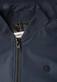 Veste bleu marine avec fermeture éclair à l'avant, col côtelé, doublure intérieure en tissu noir, et un patch logo circulaire noir sur la poitrine.