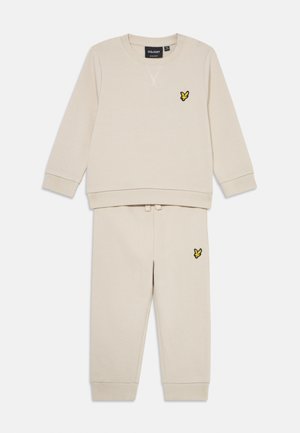 Beige toddler two-piece tracksuit with long-sleeve top and pants, featuring yellow bird logo on chest and thigh, ribbed cuffs, and snap buttons on shoulder.
