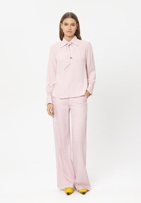 Hellrosa Bluse mit Schleifendetail, kombiniert mit passenden hellrosa ausgestellten Hosen. Gelbe spitze Schuhe mit schwarzen Akzenten vollenden den Look.