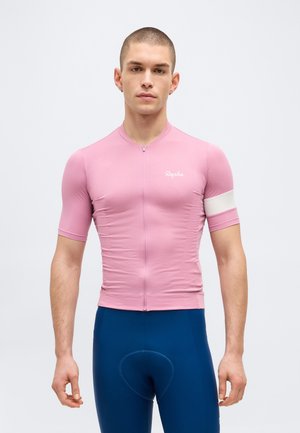 Homme portant un maillot de cyclisme rose ajusté avec un brassard blanc et un short de cyclisme rembourré bleu, debout devant un fond uni.
