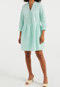Šaty v mátově zelené barvě s knoflíkovou přední částí, tříčtvrtečními rukávy a nařaseným pasem, vyrobené z texturovaného materiálu.