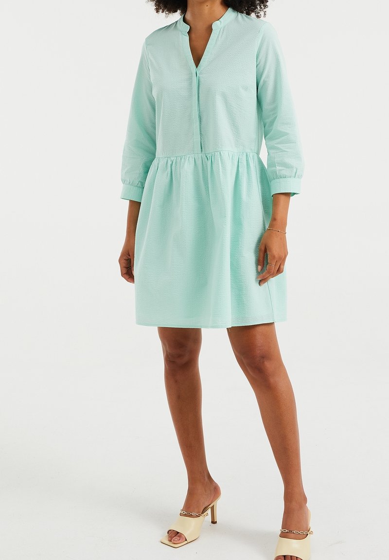 Šaty v mátově zelené barvě s knoflíkovou přední částí, tříčtvrtečními rukávy a nařaseným pasem, vyrobené z texturovaného materiálu.