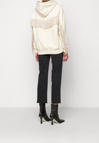 Personne vue de dos portant un sweat à capuche crème avec des détails à franges, un pantalon court noir et des bottines à talons noires sur un fond uni.