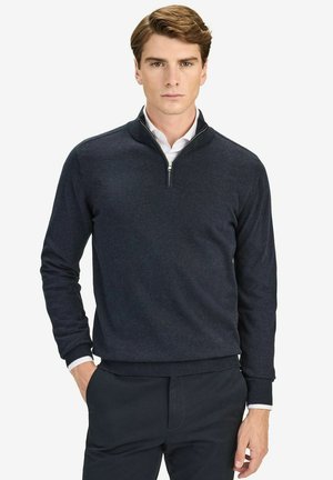 Marinblå tröja med halvdragkedja, mjuk textur och ribbade ärmslut. Stylad över en vit skjorta för kontrast. Avslappnad, figurnära design.