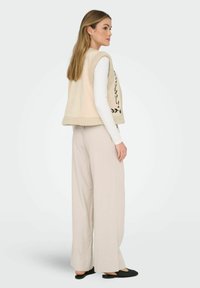 Beige ärmlös väst med en mjuk, strukturerad insida, dekorativt mönster av svarta blad, kombinerad med ljusbeiga vida byxor och svarta slip-on skor.