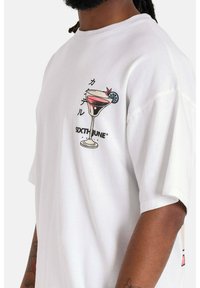 Vit t-shirt, oversized passform, med en tryckt cocktailglasdesign och texten "SIXTHJUNE" i svarta, rosa och blå detaljer.