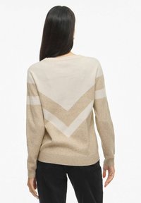 Žena s dlhými tmavými vlasmi, oblečená v béžovom a smotanovom svetri s chevronovým vzorom na chrbte a čiernych nohaviciach, stojaca na pozadí bielej steny.