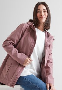 Woman wearing a mauve waterproof jacket over a white sweatshirt and blue jeans, sitting on a white surface against a plain background.