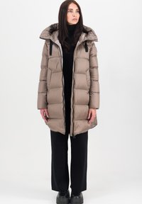 Beige gewatteerde jas met een capuchon, gestikt patroon, voorkant met rits en zwarte accenten, gecombineerd met zwarte wijde broek en platform schoenen.