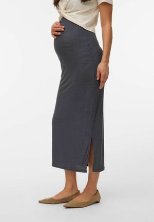 Femme enceinte portant un haut blanc, une jupe midi grise côtelée avec une fente latérale, et des ballerines beiges, debout devant un fond blanc.