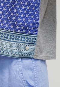 Camisa gris de manga corta con un dobladillo azul estampado con diseños intrincados y un botón a presión blanco. Tela azul clara visible debajo.