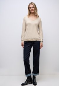 Beige svetr s výstřihem do V a dlouhými rukávy, spárovaný s tmavě modrými džíny rovného střihu s ohrnutým lemem u kotníků a černými kotníkovými botami se zlatým akcentem.