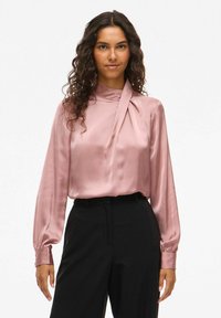 Blusa de satén en color malva con mangas largas y cuello alto; presenta un detalle anudado en el cuello y puños ajustados, combinada con pantalones negros.