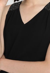 Svart ärmlös topp med V-ringning; har strukturerade, dekorerade axelaccenter. Lätt tyg med en slät finish.