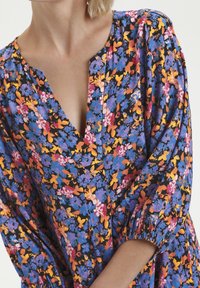 Robe à fleurs avec un fond noir, ornée de fleurs vibrantes bleues, roses, oranges et jaunes. Décolleté en V et manches bouffantes. Tissu doux.