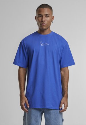 Férfi, aki élénk kék, túlméretezett pólót visel kis fehér logóval a mellkasán, és világoskék farmert, egyszerű háttér előtt áll.