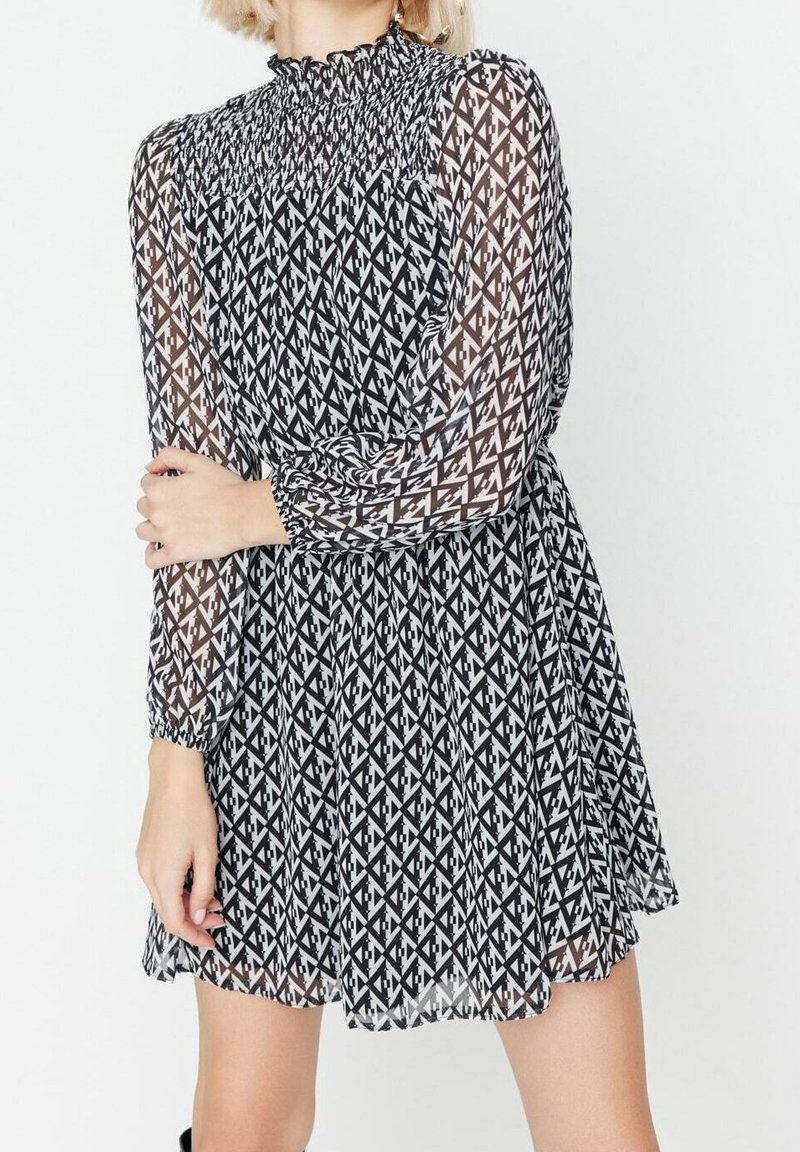 Femme portant une robe à motif géométrique noir et blanc avec des manches longues transparentes et un col smocké haut.