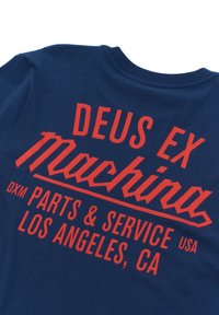 Marineblå t-shirt med fed rød typografi, der læser "DEUS EX Machina" og "DXM PARTS & SERVICE LOS ANGELES, CA." Blødt bomuldsstof.