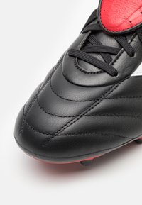 Crampons de football en cuir noir avec des accents rouges, présentant une surface texturée, un système de laçage et un design bas. La semelle a des crampons visibles.