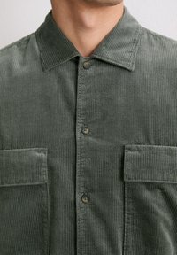 Camicia in corduroy verde con texture a costine verticali, colletto, due tasche sul petto e chiusura con bottoni.