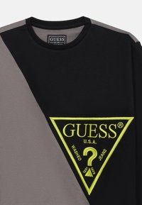 Maglietta a maniche lunghe nera e grigia con un grande logo triangolare giallo ricamato che presenta la scritta "GUESS" e un dettaglio a forma di punto interrogativo.