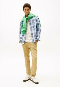 Lyseblå ternet skjorte, hvid T-shirt, grøn sweater bundet om halsen, beige bukser, og hvide sneakers med sorte og blå detaljer.